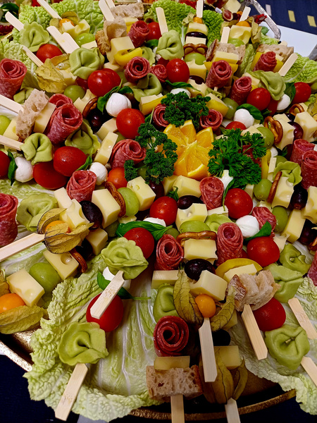 KI generiert: Das Bild zeigt eine farbenfrohe Platte mit verschiedenen Käsespießen, die mit Tomaten, Wurst, Oliven, Mozzarella und grünen Tortellini garniert sind. In der Mitte befindet sich eine dekorative Anordnung aus Orangenstücken und frischer Petersilie.