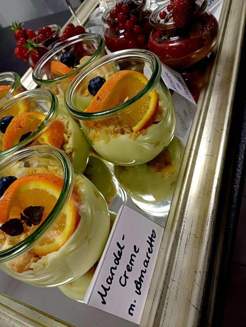 KI generiert: Ein Tablett mit Desserts in Gläsern, darunter "Mandel-Creme m. Amaretto" und Beeren-Desserts.