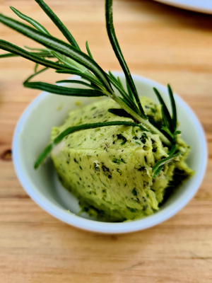 KI generiert: Das Bild zeigt einen Klecks Kräuterbutter in einem kleinen weißen Behälter, verziert mit frischem Rosmarin. Der Fokus liegt auf der cremigen Textur und den grünen Kräutern in der Butter.