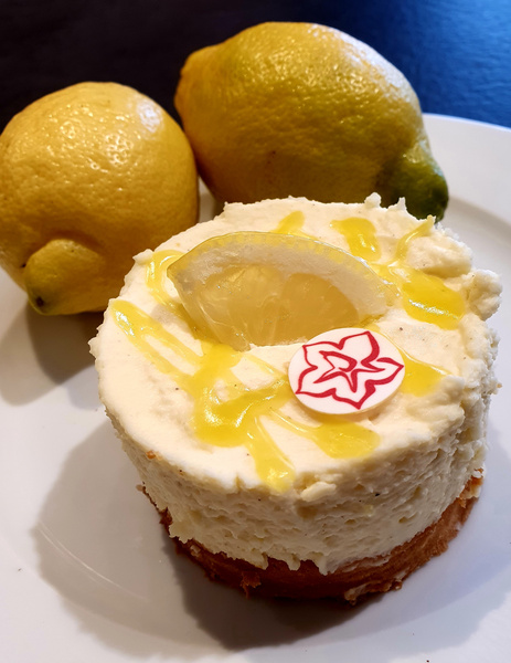 KI generiert: Das Bild zeigt einen kleinen Zitronenkuchen mit einer Zitronenscheibe und einer dekorativen Verzierung, umgeben von zwei frischen Zitronen. Der Hauptinhalt ist der Kuchen auf einem weißen Teller.