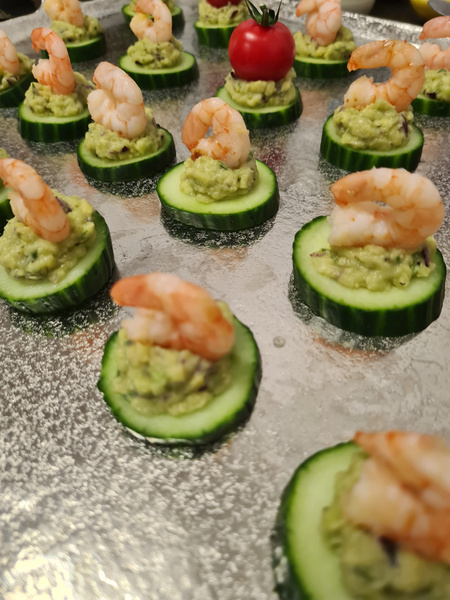 KI generiert: Das Bild zeigt eine Platte mit Appetithäppchen aus Gurkenscheiben, die mit Guacamole und Garnelen belegt sind. In der Mitte befindet sich eine Kirschtomate als Dekoration.