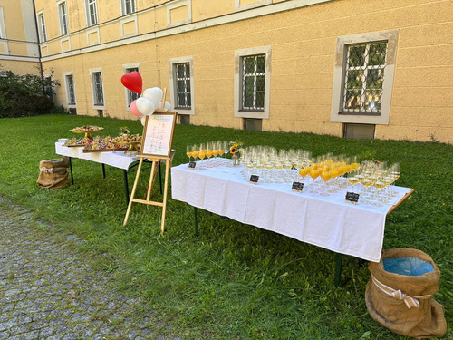 KI generiert: Ein Buffet im Freien mit Getränken und Snacks auf Tisch mit weißen Decken, daneben eine Staffelei mit Menü.