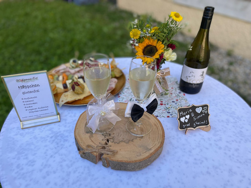 KI generiert: Ein Tisch mit Getränken, Blumen und Schildern für eine Hochzeit. Text: "Häppchen glutenfrei", "Maria & Simon grad g'heirat'".