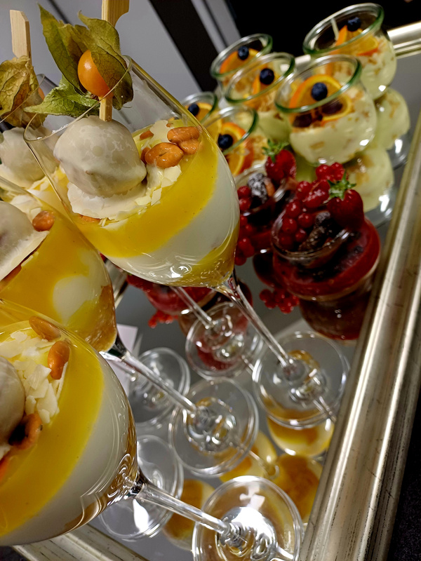 KI generiert: Das Bild zeigt eine Auswahl von kunstvoll angerichteten Desserts, darunter Gläser mit mehrschichtigen Cremes und Früchten sowie kleine Dessertgläser mit bunten Garnierungen. Die Spiegelung verstärkt die elegante Präsentation der Desserts.