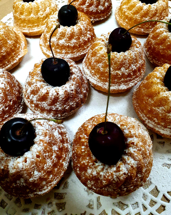 KI generiert: Das Bild zeigt eine Anordnung von kleinen Gugelhupf-Kuchen, die mit Puderzucker bestreut sind und jeweils mit einer Kirsche dekoriert wurden. Die Kuchen sind auf einer dekorativen Unterlage präsentiert.