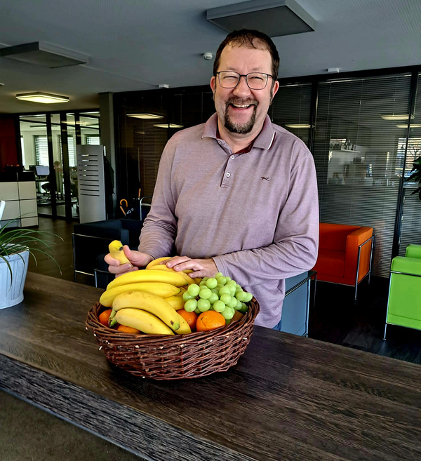 KI generiert: Ein Mann steht lachend hinter einem Obstkorb mit Bananen, Trauben und Orangen in einem modernen Büro.