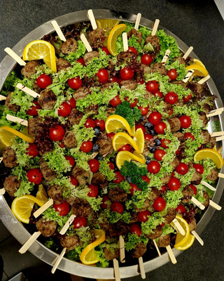 KI generiert: Das Bild zeigt eine große Platte mit Spießen, die Fleischbällchen, Kirschtomaten und Salatblätter enthalten, dekoriert mit Orangenscheiben und Heidelbeeren. Die Anordnung der Speisen ist kreisförmig und ansprechend angerichtet.