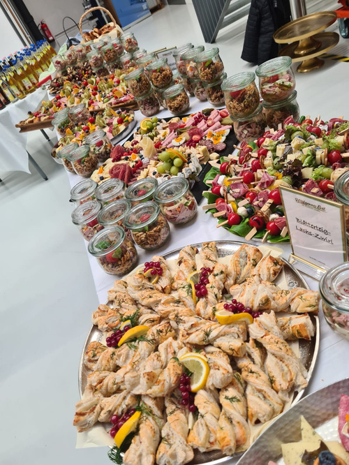 KI generiert: Ein Buffet mit vielfältigen Speisen, darunter Salate in Gläsern, Früchte und Gebäck. Text: "Blätterteig Lachs-Zwirbel".